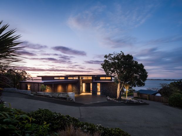 Twilight exterior view of a low, modern single-storey residence on a sloping site. The house features dark horizontal cladding, warm timber-lined areas, large glazed openings, a flat roof with clerestory-like windows, and a paved driveway. Landscaping and mature trees frame the property, with a body of water visible in the distance.