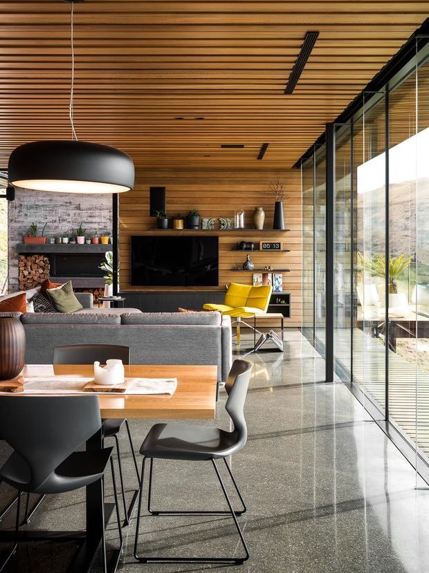 A refined open-plan living and dining area with extensive floor-to-ceiling glazing, a warm timber slatted ceiling, horizontal timber wall panelling, polished concrete flooring, and dark-framed glass doors. The room includes a contemporary dining table with black chairs, a grey sofa, a bright yellow lounge chair, built-in shelving with firewood and decor, and a wall-mounted television. Strong natural light and views through the glazing create an airy connection to the outdoors.