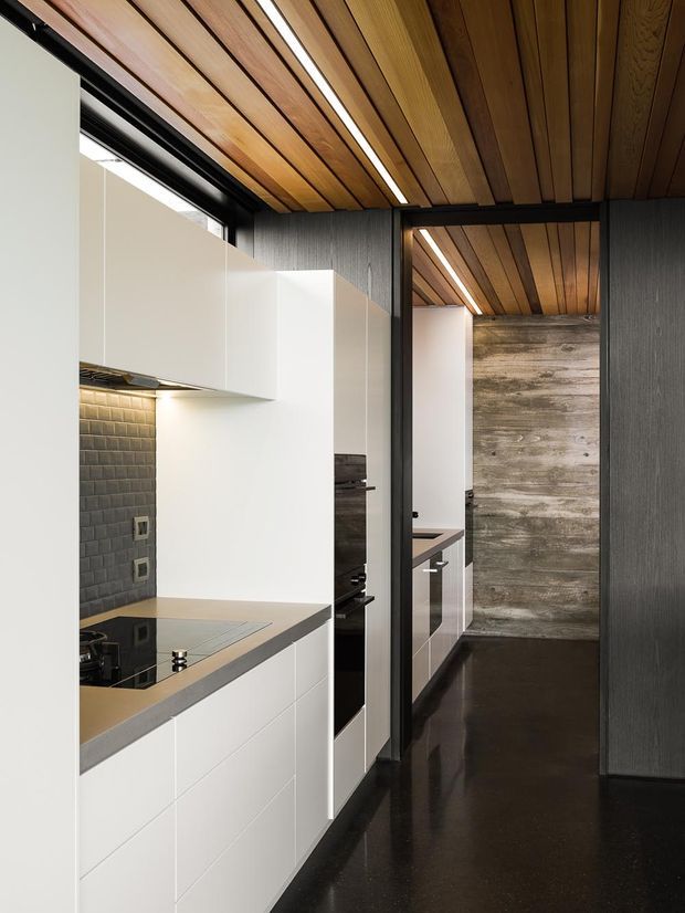 A narrow, contemporary galley kitchen with crisp white flat-panel cabinetry, a grey stone benchtop, dark textured tiled splashback, and integrated black appliances. The kitchen opens toward a dark polished corridor with a textured timber feature wall. Warm timber ceiling battens with linear recessed lighting extend through the space, contrasting with charcoal wall finishes and flooring.