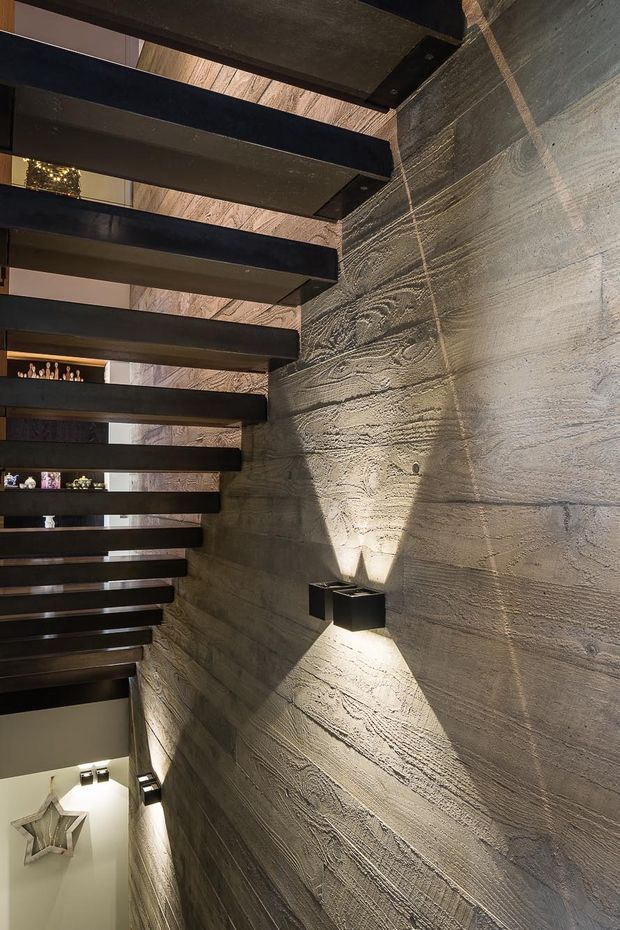Interior view of a dramatic modern stairway with open-riser black metal or dark timber treads spanning alongside a heavily textured concrete wall. Integrated wall sconces cast upward and downward beams across the rough surface. A lower-level area and decorative shelving are partially visible beneath the stairs, creating a strong sculptural, industrial atmosphere.