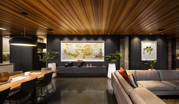 A sophisticated open-plan living and dining area with a dramatic dark feature wall, low built-in cabinetry, large framed artwork, and a substantial sectional sofa. The ceiling is clad in warm timber battens with integrated recessed lighting, while a sculptural black pendant hangs above the timber dining table. Polished dark flooring, indoor palms, and neutral furnishings create a refined, moody atmosphere.