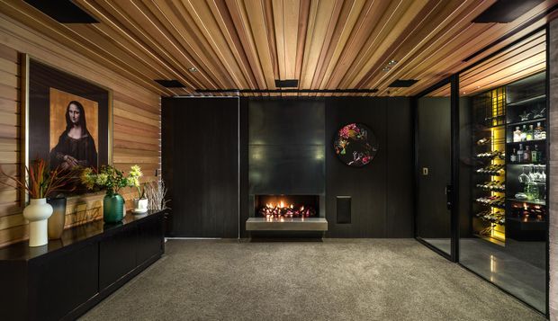 A dark, sophisticated media or living room with extensive timber wall and ceiling lining, a black feature fireplace wall with a linear fire, and a built-in glass-fronted bar or drinks display on the right. Black low cabinetry runs along the left wall beneath artwork and decorative plants. The room has a moody, luxurious atmosphere, with recessed and concealed lighting highlighting the timber ceiling and dark finishes.
