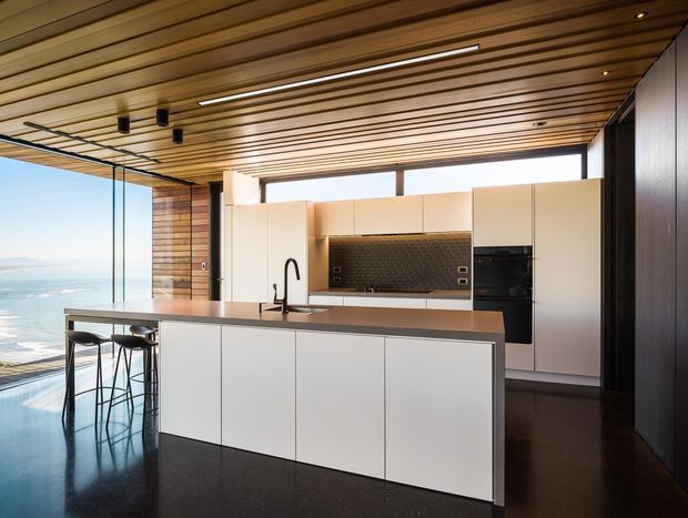 A contemporary open-plan kitchen with a large white island, dark stone-look benchtop, integrated sink and black tap, tall white cabinetry, dark tiled splashback, and built-in black appliances. Warm timber battens line the ceiling and part of the walls, while full-height glazing opens to a visible ocean and beach outlook. Dark polished flooring and black-framed glazing create a refined, high-contrast interior.