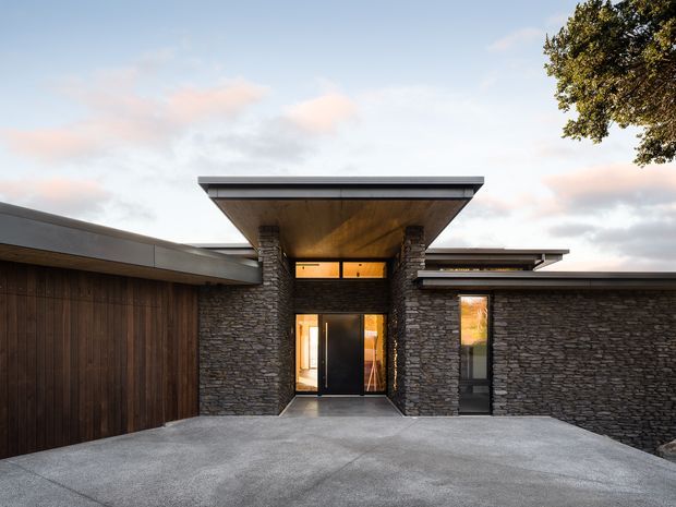 Front exterior view of a low, modern residence with a strong horizontal composition. The entrance is framed by rough stacked-stone walls and columns beneath a broad projecting timber-lined canopy, with dark timber garage doors and warm-toned glazing. A wide concrete driveway occupies the foreground.