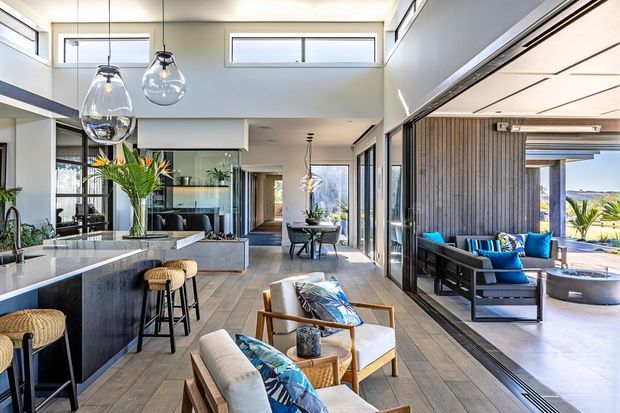 Bright, spacious open-plan contemporary interior with a kitchen island in the foreground, dining and living areas extending toward the rear, and a connected covered outdoor lounge visible through full-height sliding glazing. The room features a double-height void, clerestory windows, large pendant lights, pale tiled flooring, dark kitchen cabinetry, timber details, and blue accent furnishings. The outdoor area includes a lounge setting and views toward landscaped surroundings.