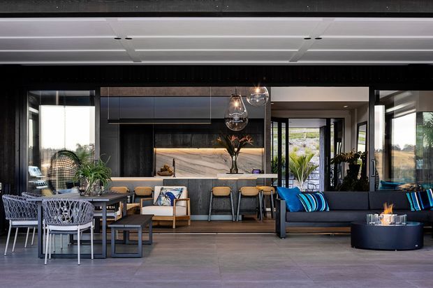 A sophisticated open-plan indoor-outdoor living and entertaining area with a contemporary kitchen and island bench, dining table, bar seating, and a dark sectional lounge. Extensive black-framed glazing opens toward an outdoor setting, while layered pendant lights, dark cabinetry, timber accents, and blue cushions create a refined modern atmosphere.