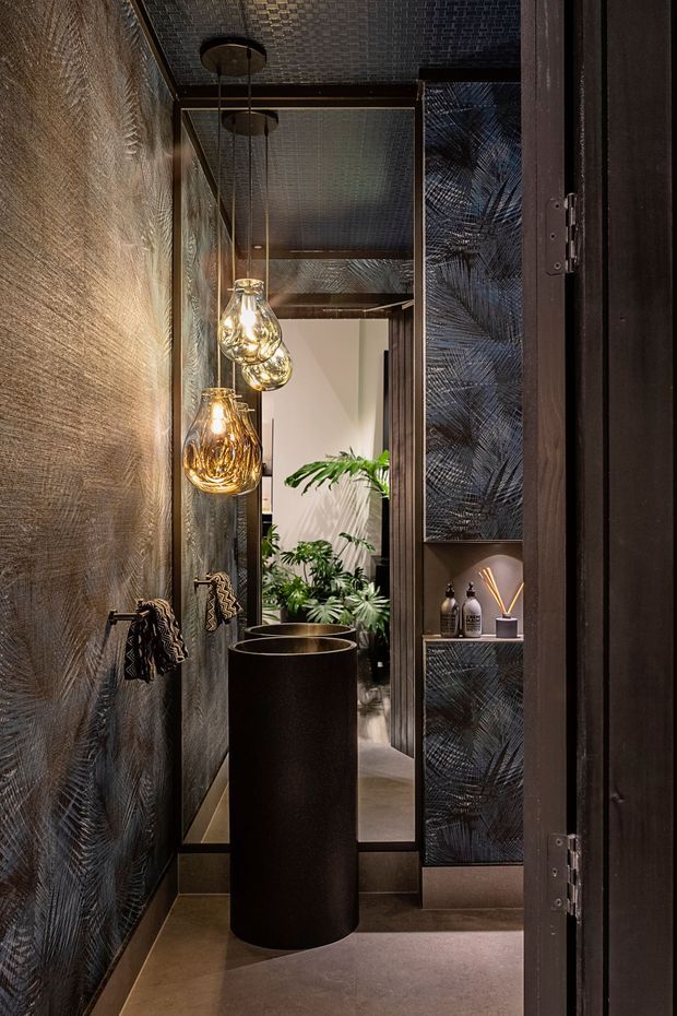 A narrow, darkly styled bathroom or powder room viewed through an open door, featuring a tall round black vessel basin, wall-mounted black taps, textured dark wall finishes, mirrored cabinetry, hanging glass pendant lights, and a small indoor plant. The space has a refined, dramatic, boutique-hotel atmosphere with metallic and black accents.