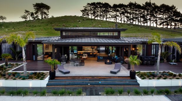 A wide exterior view of a low, modern pavilion-style residence set into a landscaped hillside. The dark metal roof and extensive floor-to-ceiling glazing frame an open entertaining terrace with outdoor seating, potted palms, steps, and a long narrow reflecting or swimming pool in the foreground. A tree-covered rural landscape rises behind the house.