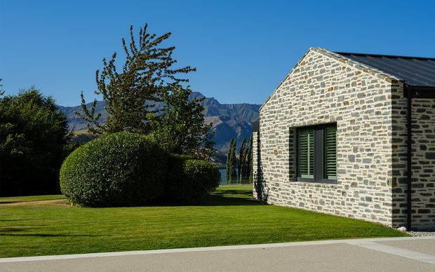 Exterior view of a low, gabled stone building with a dark metal pitched roof and dark green shuttered window. The building sits beside a manicured lawn and clipped hedges, with dramatic mountain scenery visible beyond.