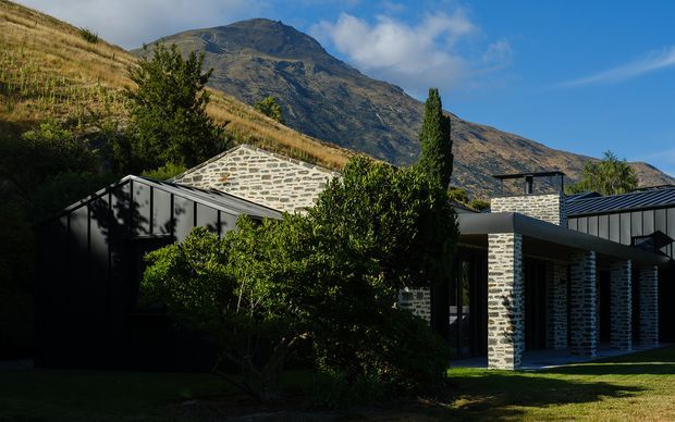 Exterior view of a contemporary hillside residence set against a dramatic mountain landscape. The home combines dark metal-clad roof and wall sections with pale grey stacked-stone masonry, deep roof overhangs, and large areas of glazing, partially screened by mature planting and lawn.