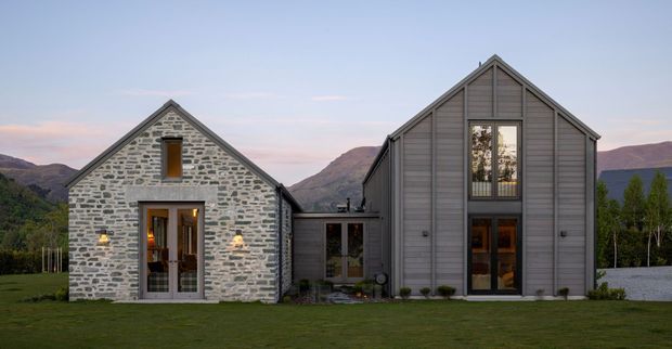 Exterior view of a contemporary gabled home composed of two connected volumes. The left wing has a light grey irregular stone facade with a steep gable and dark-framed glazed entry, while the taller right wing is clad in vertical grey weatherboards with large multi-level windows. A lower glazed connector joins the wings. The house sits in a landscaped lawn with mountains visible in the background at dusk.