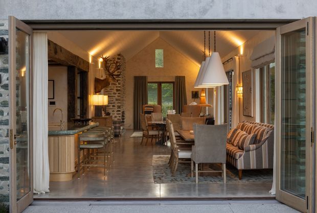 View through wide-open glazed folding doors into a compact, warmly lit open-plan living and kitchen area. The room has a vaulted timber-lined ceiling, exposed rough stone and timber architectural elements, polished concrete flooring, a compact kitchen with a stone benchtop and fluted cabinetry, dining furniture, upholstered seating, and layered pendant and wall lighting. The opening connects directly to an exterior terrace or courtyard.