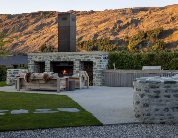 A landscaped outdoor entertaining area with a built-in stone fireplace and tall chimney, positioned against a dramatic alpine mountain backdrop. A timber-framed outdoor sofa with throws faces the fire, while a paved terrace, clipped lawn, gravel edging, stone walls, and a partially visible outdoor kitchen or bar complete the setting.