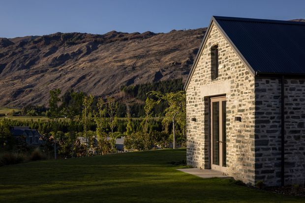 Exterior view of a stone-clad gable-roof building set beside a broad lawn, with rugged mountain terrain and landscaped vegetation in the background. The building has pale coursed stone walls, dark roofing, a tall glazed timber-framed door, and a narrow vertical window opening.