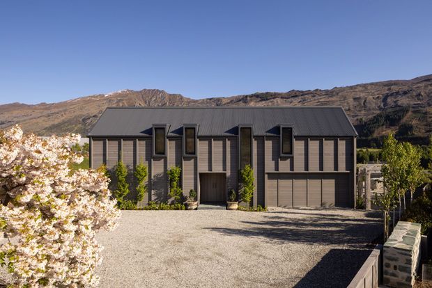 A contemporary multi-level residential house set against a rugged mountain landscape. The dark grey gabled roof and matching vertical cladding create a restrained facade, with tall narrow windows, a central recessed entry, an integrated double garage, and a broad gravel driveway. Flowering and leafy trees frame the foreground, while stone retaining walls edge the property.