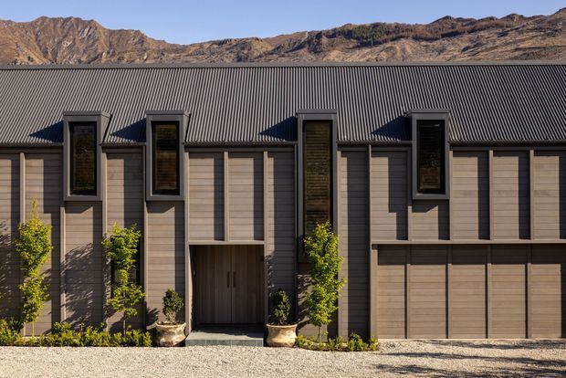 A modern residential facade set against rugged rocky hills. The building has a long, symmetrical composition with dark corrugated metal roofing, vertically arranged dark-framed windows, warm grey-brown cladding, a recessed entry, and an integrated double garage. Young ornamental trees and gravel landscaping line the frontage.