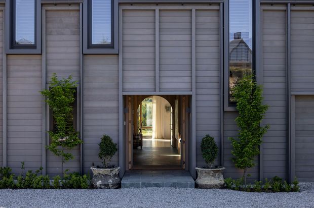 Front facade of a refined contemporary residence with symmetrical grey vertical and horizontal cladding, tall narrow dark-framed windows, and a timber-framed arched entry opening. Climbing greenery and formal planters flank the doorway, with a gravel forecourt. The entry offers a view through to a bright interior hall.