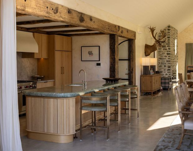 A warm, open-plan kitchen and dining area in a rustic renovated interior. The kitchen features timber cabinetry, a large rounded island with a blue-green stone benchtop, integrated sink and bar seating. Exposed heavy timber beams frame the opening, while a rough stone chimney or feature wall, polished concrete floor, vintage furniture, and mounted antlers contribute to a rustic character.