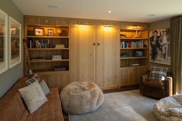 Warm, intimate living room with extensive custom timber built-in cabinetry, bookshelves, display niches, and central double doors. The room includes a tufted brown leather sofa, leather armchair, oversized textured ottoman, pale area rug, framed artwork, and layered warm shelf lighting. The styling is rustic and refined, with muted green-grey walls and dark timber flooring.