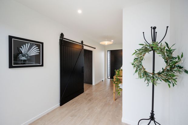 Bright contemporary residential entry hall with white walls, pale timber flooring, a black sliding barn-style door, framed monochrome artwork, and a decorative black coat stand with a leafy wreath. A small console or table with greenery is visible deeper in the hallway.