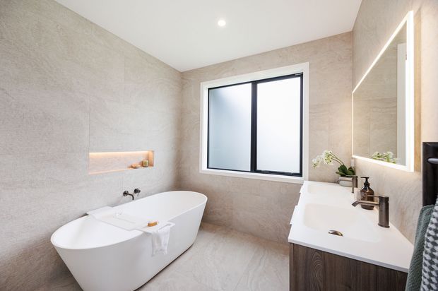 Bright contemporary bathroom with large-format light grey stone-look wall and floor finishes, a freestanding white bathtub, double vanity with dark timber cabinetry, black tapware, a framed window, recessed illuminated niche, and a large backlit mirror.