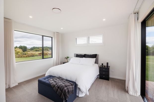 Bright, contemporary bedroom with a neatly made bed, dark upholstered headboard, black bedside table, and a navy upholstered bench at the foot of the bed. The room has soft grey carpeting, white walls and ceiling, full-height curtains, recessed lighting, a wide horizontal window, and a glazed door opening toward an outdoor area. The neutral palette and abundant daylight create a clean, calm atmosphere.