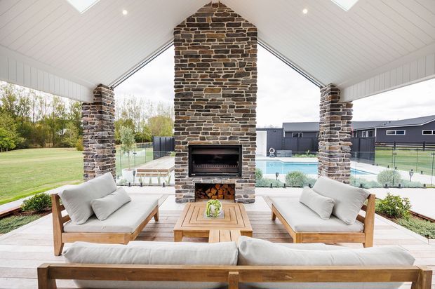 A bright, covered outdoor living area with a vaulted white-paneled ceiling, exposed stone fireplace chimney and built-in fireplace, timber-framed upholstered seating, and a timber coffee table. The open sides overlook a landscaped lawn and swimming pool, with a neighboring dark-clad building beyond.