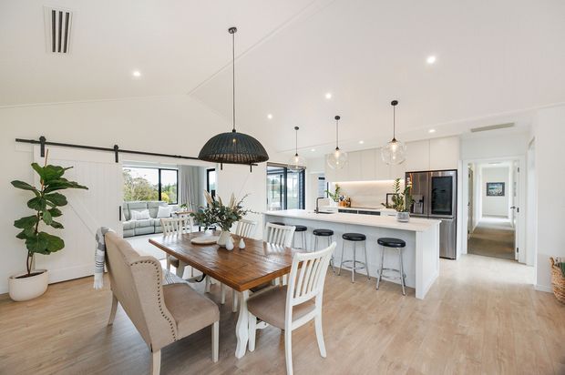 Bright open-plan residential interior combining a dining area, kitchen and living space. The room features white walls and cabinetry, light timber flooring, a large island with bar seating, pendant lighting, a timber dining table with mixed seating, and a black barn-style sliding door track. Glazing opens toward an outdoor area, while a hallway is visible beyond the kitchen.