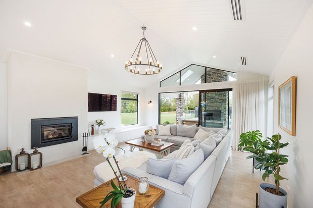 Bright, spacious contemporary living room with a large light-coloured sectional sofa arranged around a timber coffee table and a built-in fireplace. The room features white walls and a white-lined ceiling, pale timber flooring, expansive black-framed glazing, a high clerestory-style window, sheer curtains, recessed lighting, and a prominent suspended metal pendant. Indoor plants and neutral furnishings create an airy, refined atmosphere.