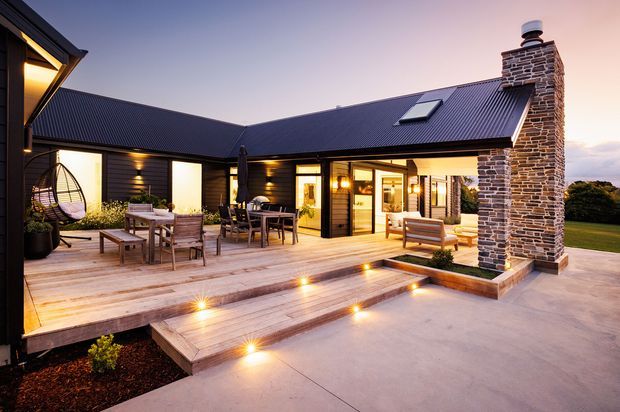 Twilight exterior view of a modern single-storey residence arranged around a covered timber deck and patio. The home has dark horizontal cladding, a steep dark metal gable roof with skylight, and a prominent stacked natural-stone chimney. Warm interior and recessed deck lighting illuminate the outdoor dining and lounge areas, with broad timber steps leading to a concrete terrace.