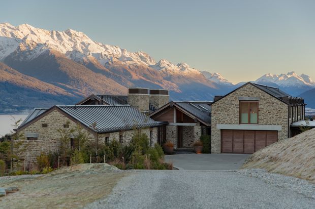 Exterior view of a substantial mountain house built from pale textured brick and dark standing-seam metal roofing. Multiple pitched roof volumes, exposed timber gable detailing, stone or brick chimney stacks, and a double garage frame a gravel driveway. Snow-capped alpine mountains and a lake are visible behind the property.