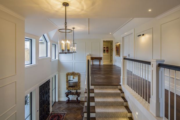 Elegant traditional-style residential stair hall and landing with a patterned carpet runner, dark herringbone timber flooring, white wall panelling and trim, black stair balusters and handrails, and tall multi-pane windows. A tiered chandelier and wall sconces provide warm lighting, while a console table and ornate mirror sit beneath the landing.