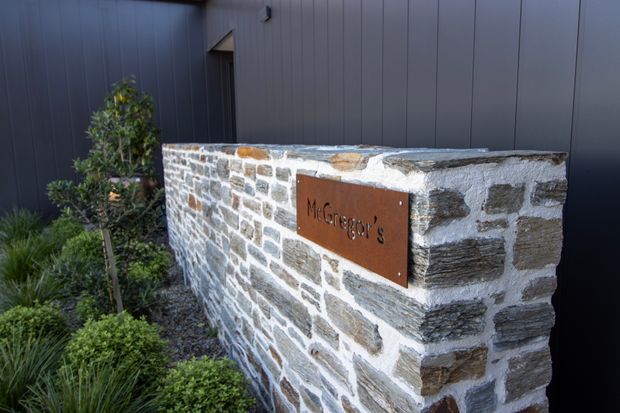 Exterior entrance feature with a low, rough-stone masonry wall bearing a rust-toned metal plaque reading “McGregor’s.” The wall sits beside landscaped planting and contrasts with a dark vertical-panel building facade and a partially visible doorway. The image emphasizes the textured stonework and property identification sign.