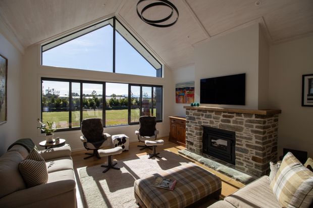 A bright, spacious living room with a vaulted timber-lined ceiling, expansive corner glazing, and views over a landscaped rural setting. The room features a substantial natural stone fireplace with a timber mantel, wall-mounted television, upholstered sofas, two lounge chairs, and a large plaid ottoman arranged on a light rug.