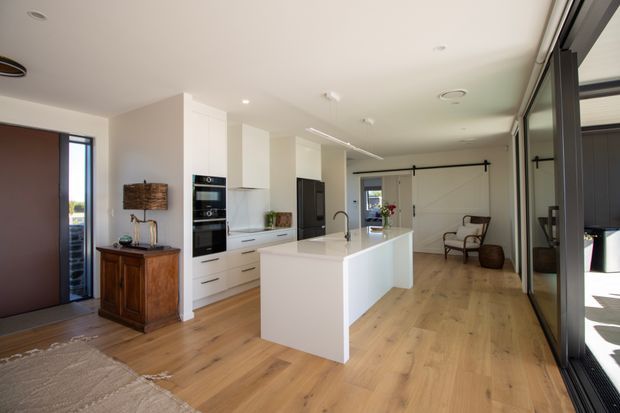 Bright, open-plan contemporary kitchen and living area with white built-in cabinetry, integrated black appliances, a large white waterfall island with sink, and light timber flooring. Full-height dark-framed glazing and sliding doors line the right side, while a lounge area with an armchair is visible beyond the kitchen. A timber console and woven lamp add warmth to the otherwise clean, minimal interior.