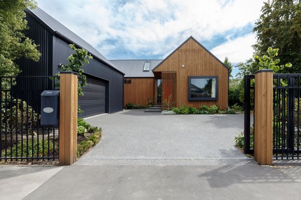 A contemporary single-storey home viewed from the street through an open black metal gate. The property features a gabled timber-clad front volume, a dark vertically ribbed garage wing with a broad roller door, dark metal pitched roofing, and a paved driveway bordered by low planting. Timber gate posts and black metal fencing frame the entrance.