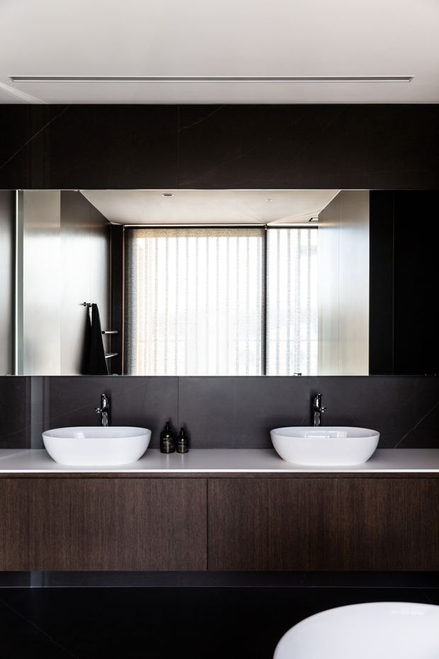 A contemporary double vanity bathroom featuring two white vessel basins on a pale stone countertop, dark timber cabinetry, black wall and floor finishes, and black tapware. A large mirrored wall reflects a bright window with sheer blinds, while recessed ceiling lighting and a linear air vent add to the streamlined design.