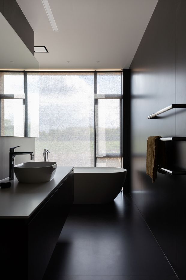 A sophisticated contemporary bathroom featuring a freestanding white bathtub, sculptural vessel basin on a dark vanity, black wall and floor finishes, and sleek wall-mounted fixtures. Full-height translucent glazing provides soft natural light and privacy, creating a dramatic, minimalist atmosphere.