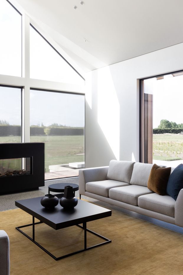 A bright, minimalist living room with expansive floor-to-ceiling glazing overlooking open farmland. The space features a light grey upholstered sofa with contrasting brown and navy cushions, a low black rectangular coffee table with sculptural black vessels, a black-framed fireplace, and a warm tan rug. White walls and ceiling create a clean, airy interior with strong natural light.