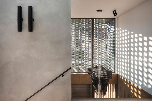 A refined stair landing with a minimalist concrete-look plaster wall, black cylindrical wall lights, and a slim black handrail. Perforated breeze-block screening wraps the stairwell glazing, casting a strong geometric pattern of light and shadow across the white wall. Warm timber built-in joinery and a sculptural woven pendant add contrast.