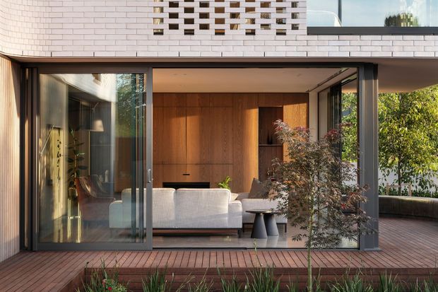 A contemporary residential living space opening onto a timber deck through expansive sliding glass doors. The room features warm timber wall panelling and built-in joinery, a light upholstered sofa, dark coffee tables, and a sheltered outdoor courtyard with planting. The upper facade is clad in pale brick with a perforated brick screen, while the interior and deck are framed in dark metal.