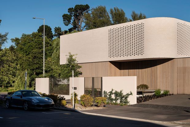 Street-facing view of a contemporary multi-level building with a curved pale rendered upper volume featuring perforated brick screens, a warm vertical timber-clad lower level, recessed glazing, landscaped frontage, and a paved driveway. The image appears to show a commercial or institutional architectural project in a leafy suburban setting.