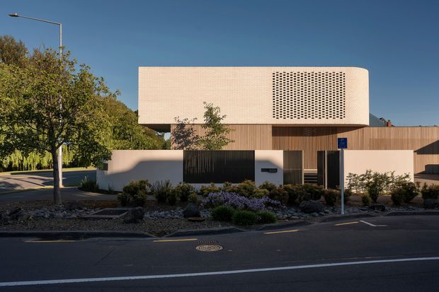 Street-facing view of a contemporary low-rise building with a warm timber-clad ground level, pale rendered upper volume, and a distinctive perforated breeze-block screen. The building is set behind landscaped planting and a low boundary wall along a paved urban street, with a clean geometric facade and flat roofline.