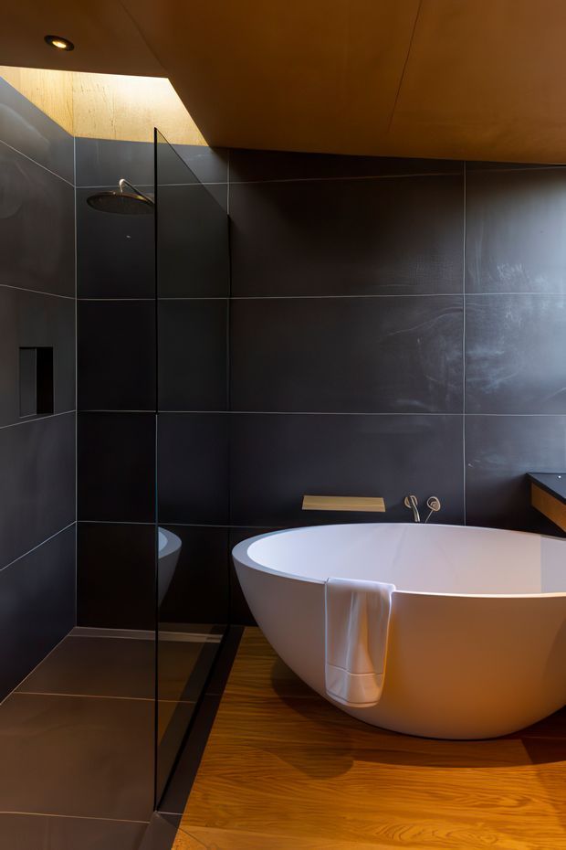 A dramatic, contemporary bathroom featuring a freestanding white oval bathtub beside a frameless glass shower screen. Large charcoal-black wall and floor tiles create a dark, monolithic backdrop, while a warm timber floor and ceiling add contrast. A narrow skylight or clerestory opening casts natural light across the shower area, complemented by recessed ceiling lighting and understated brass-toned fixtures.