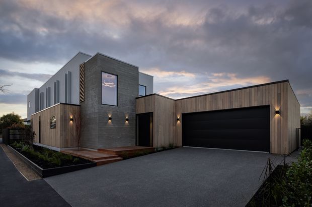 Contemporary multi-level residential house photographed from the street at dusk. The façade combines dark grey masonry, vertical timber cladding, white upper wall sections, large dark-framed windows, and a broad integrated double garage. A paved driveway, low landscaped garden beds, exterior wall lighting, and a recessed entry complete the composition beneath a dramatic cloudy sky.