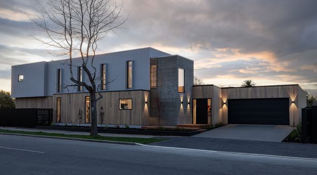 A street-facing photograph of a contemporary multi-level residence at dusk. The house combines a smooth light-grey upper volume with darker timber-look vertical cladding at ground level, a charcoal textured feature wall, narrow vertical windows, and a prominent integrated double garage. A paved driveway and front path lead to the recessed entry, while a leafless street tree partially frames the facade.