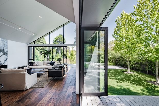 Bright contemporary indoor-outdoor living area with extensive floor-to-ceiling glazing and a fully opened glazed door connecting the room to a timber deck and landscaped lawn. The interior features a polished white ceiling, dark-framed windows, exposed dark timber flooring, and a neutral seating arrangement around a low coffee table. Mature trees and a manicured garden are visible outside.