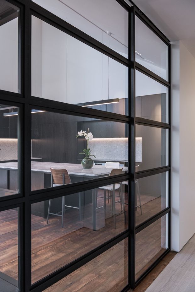 Interior view through a black-framed glazed partition into a sophisticated kitchen and dining area. The space features dark full-height cabinetry, a pale stone island with bar seating, integrated linear pendant lighting, a textured illuminated backsplash, a timber floor, and restrained contemporary styling. The black steel-look framed glazing strongly separates the foreground circulation area from the kitchen.
