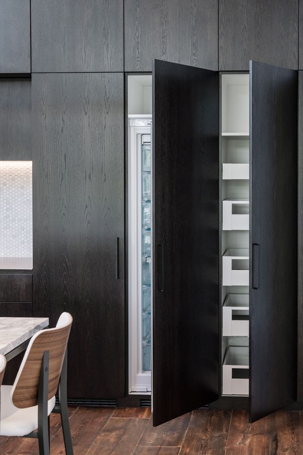 Close-up interior view of a dark wood kitchen wall with floor-to-ceiling integrated cabinetry. Tall cabinet doors are open to reveal a built-in refrigerator and narrow white shelving drawers, while part of a dining table and chairs is visible at the left. The space has dark timber flooring and a refined, minimalist appearance.