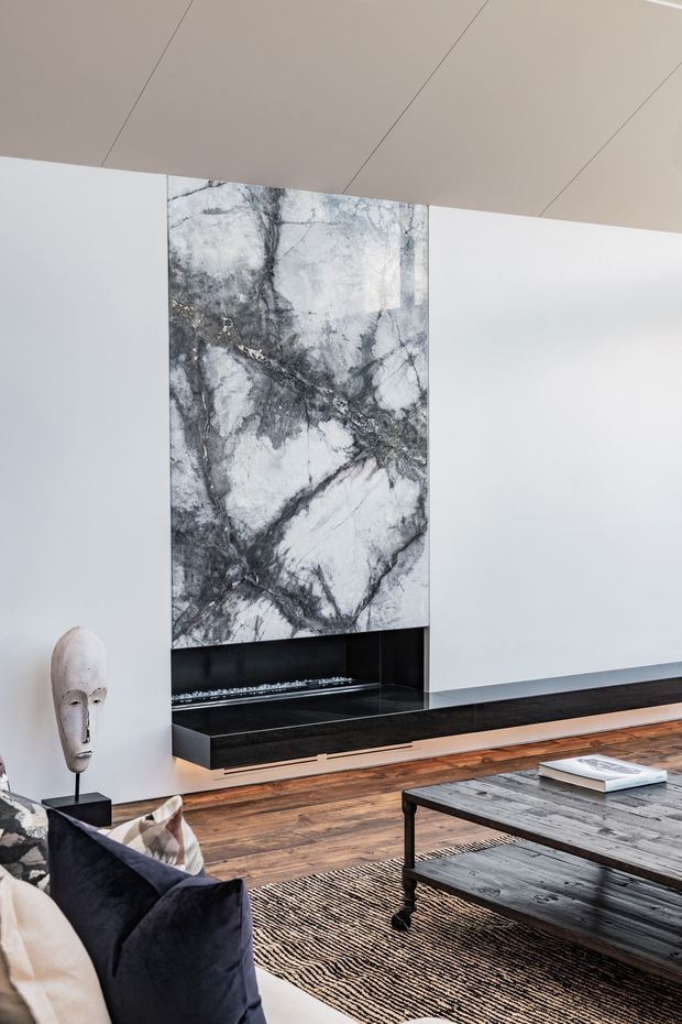 A refined contemporary living room featuring a dramatic veined marble fireplace surround and low black hearth set into white walls. Warm timber flooring, a dark upholstered sofa, patterned rug, sculptural decorative object, and rustic dark timber coffee table create contrast. The ceiling has clean linear panel joints and the room is softly lit with a minimalist, high-end aesthetic.